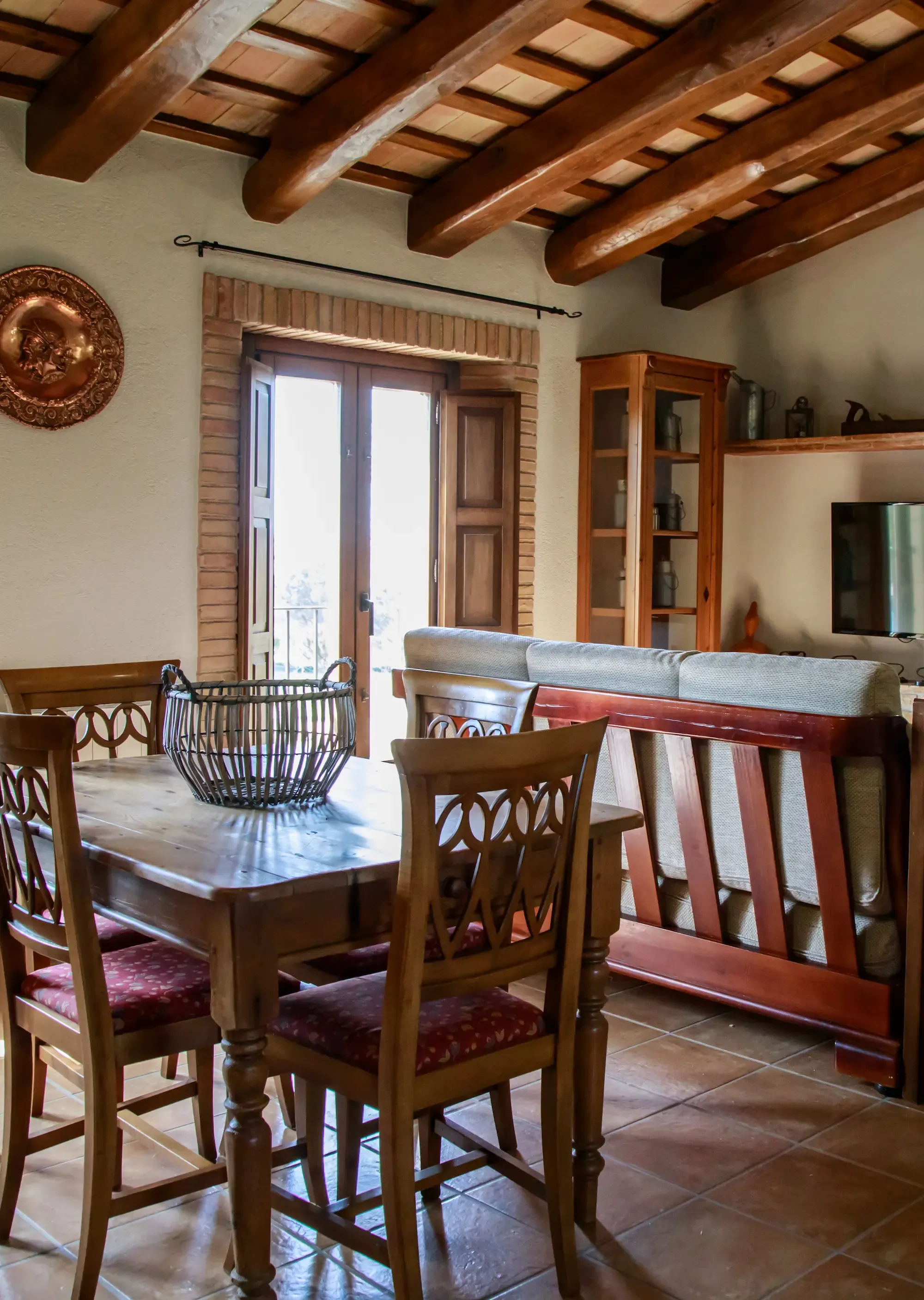Una zona de viure i menjar rústica amb vigues de fusta, rajoles de terracota, una taula de menjador de fusta, un sofà i un televisor de pantalla plana.